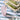 A clear acrylic drinking cup with an abstract vertical pattern in white, shown full of a light pink drink and steel bent straw. Displayed on a checkered picnic rug with a stack of 3 storage containers filled with cut vegetables.