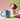 A group of 3 matte ceramic mugs with braided handles, shown on a multi coloured background. The terracotta mug is shown resting inside the yellow mug with the light blue coloured mug in the foreground.